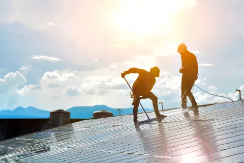 Zwei Arbeiter putzen Photovoltaik-Module auf einem Dach mit Sonne im Hintergrund.